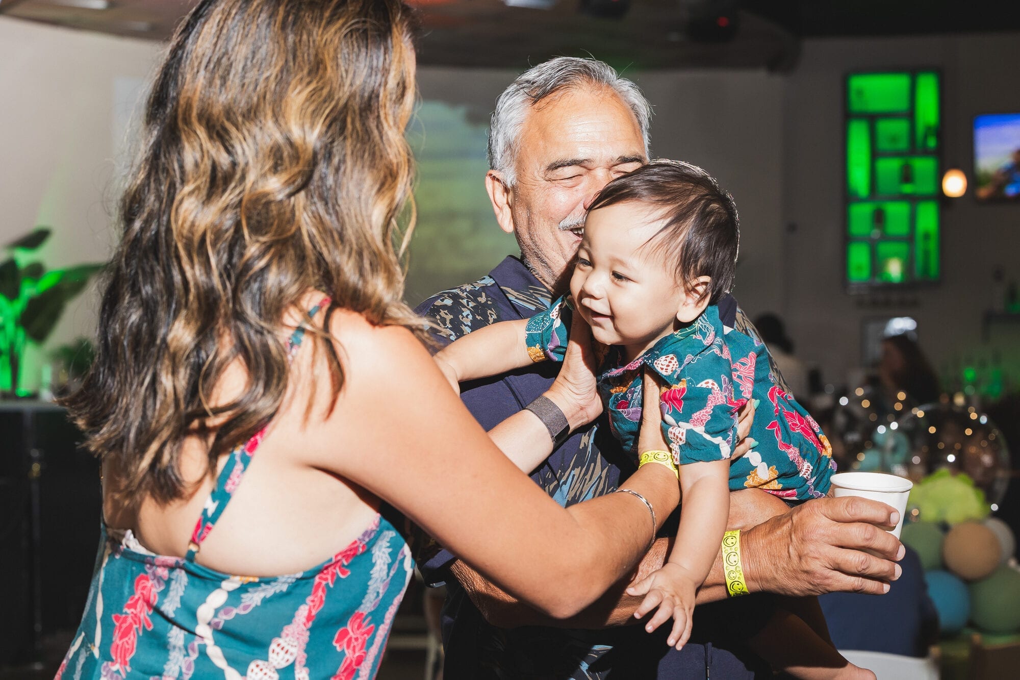 Grandfather holds a smiling baby in matching aloha shirts at a Hawaii first-birthday celebration