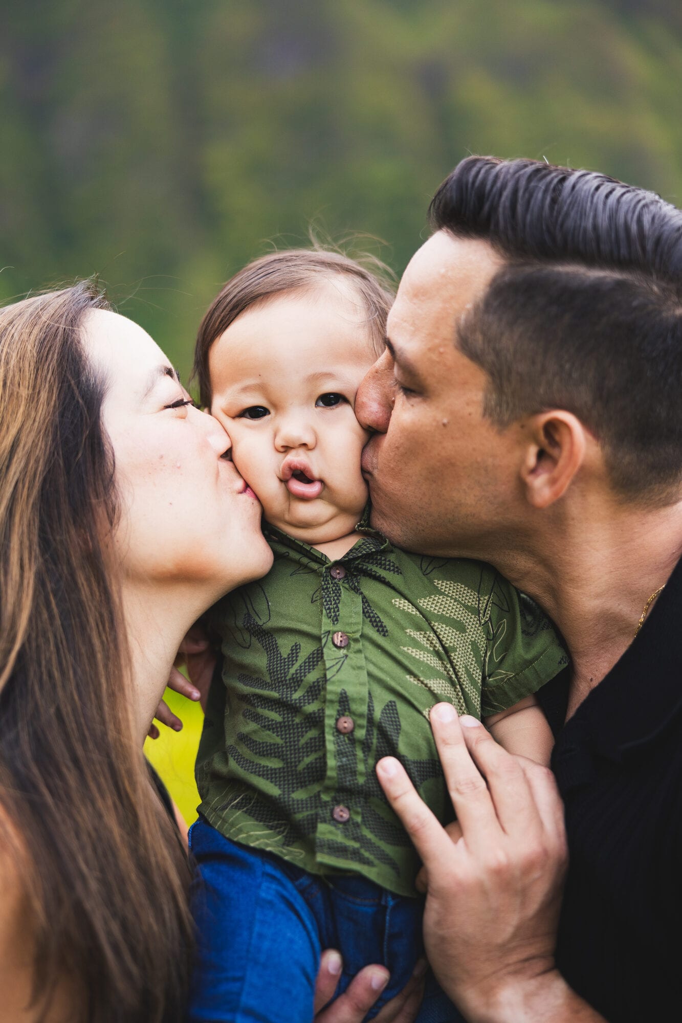 Parents kiss their baby's cheeks during a family session with a lush Oahu mountain backdrop