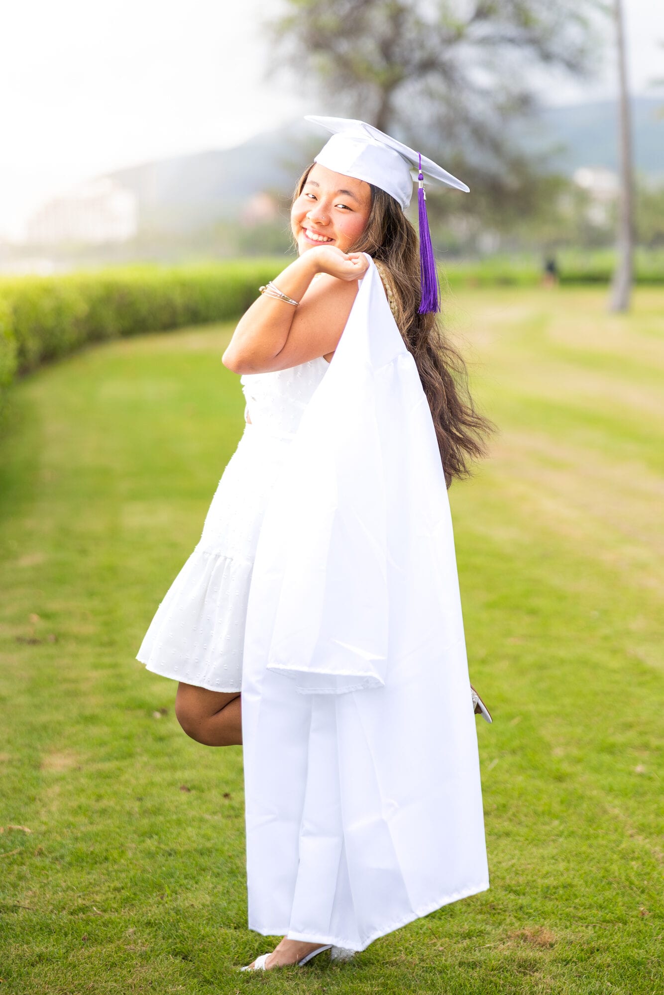 Smiling graduate in white cap and gown posing outdoors at a scenic Oahu park