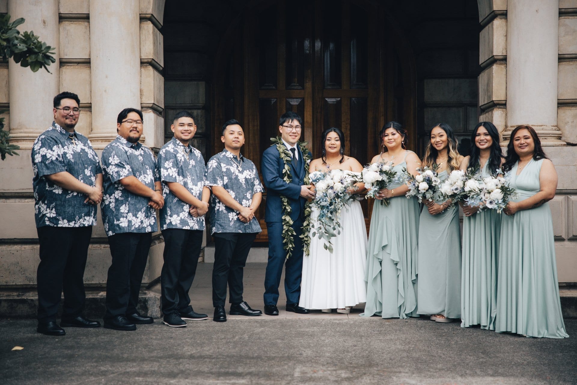 Full wedding party portrait with groomsmen in aloha shirts and bridesmaids in sage green at historic Honolulu venue